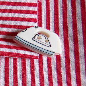 May include: Close-up of red and white striped fabric with a small, white, iron-shaped button. The button has a black and blue outline and a small iron graphic. The fabric is folded over.