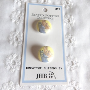 May include: Two round buttons from the Beatrix Potter Collection, featuring a bunny illustration on a yellow background. The buttons are attached to a white card with blue borders and the text "CREATIVE BUTTONS BY JHB".