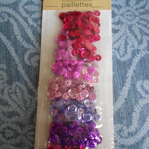 May include: A package of six bags of assorted sequins in shades of pink, purple, and blue. The sequins are arranged in a gradient pattern, with the pink sequins at the top and the blue sequins at the bottom. The package is labeled "sequins paillettes" and "6 bags sacs".