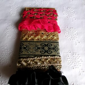 May include: A stack of decorative ribbons in various colors and textures. The top ribbon is gold with red accents, followed by a bright pink lace ribbon. Below are gold and black patterned ribbons, and a black ruffled ribbon at the bottom.