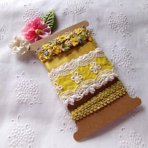 May include: A brown cardboard spool holds several spools of decorative ribbon. The ribbons are yellow, white, and green, with floral and lace designs. A pink and white flower and green leaf accent the top of the spool. The background is white fabric with floral embroidery.