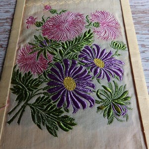 May include: A vintage embroidered fabric panel featuring a bouquet of pink and purple flowers with green leaves. The flowers are stitched in a detailed, realistic style.