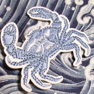 May include: A blue crab patch with intricate embroidered details. The crab is light blue with a white outline, set against a blue and white wave pattern background. This patch is suitable for clothing or accessories.