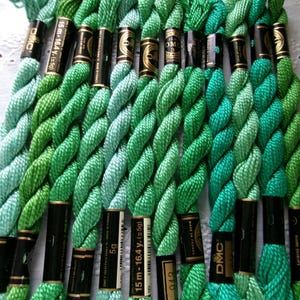 May include: A collection of DMC embroidery floss skeins in various shades of green, ranging from pale mint to deep forest green. Each twisted cotton thread bundle features a black paper label with gold text indicating 15m-16.4y and 5g.