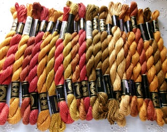 May include: A collection of DMC embroidery floss skeins arranged in a row, featuring a warm color palette of reds, oranges, golds, and browns. Each skein is secured with a black paper label displaying the brand name and product details.
