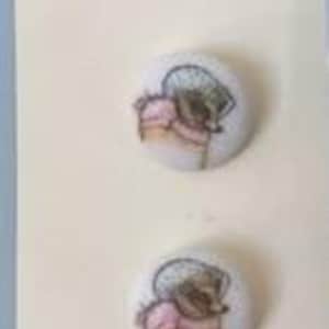 May include: Two round buttons from the Beatrix Potter Collection. Each button features a watercolor illustration of a character wearing a bonnet and pink clothing. The buttons are on a cream-colored card.