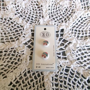 May include: Two round buttons with a Peter Rabbit design on a white background. The buttons are attached to a white and blue card with the text "JHB International" and "Made in Japan". The background is a white lace doily.