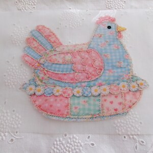 May include: A fabric applique of a chicken in shades of pink, blue, and white. The chicken has a patchwork design with floral and heart patterns. The base is a patchwork of pink and blue squares, and the edges are trimmed with small white daisies.