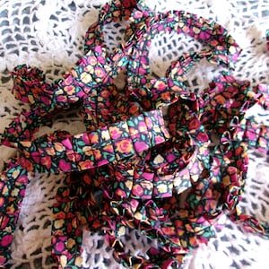 May include: Close-up of floral patterned ribbon in shades of pink, yellow, and green on a black background. The ribbon is ruffled and appears to be made of fabric. The background is a white, crocheted lace.