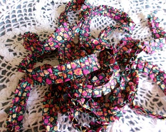 May include: Close-up of floral patterned ribbon in shades of pink, yellow, and green on a black background. The ribbon is ruffled and appears to be made of fabric. The background is a white, crocheted lace.