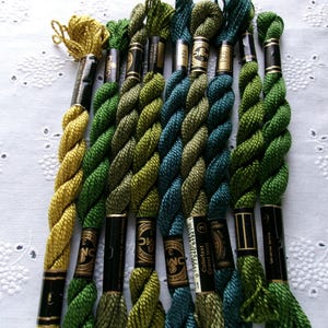 May include: A collection of eight skeins of embroidery floss in various shades of green, yellow-green, and teal. Each skein is twisted and secured with a black paper label featuring gold text and branding, arranged vertically in a parallel row.
