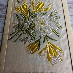 May include: A framed piece of fabric with an embroidered design of yellow and white flowers. The flowers are arranged in a bouquet and are surrounded by green leaves.
