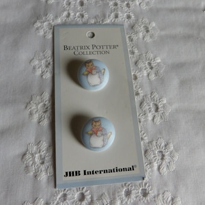 May include: Two blue fabric buttons with a white background featuring a Beatrix Potter character, Peter Rabbit, in a blue dress. The buttons are packaged on a white card with the text "Beatrix Potter Collection" and "JHB International".