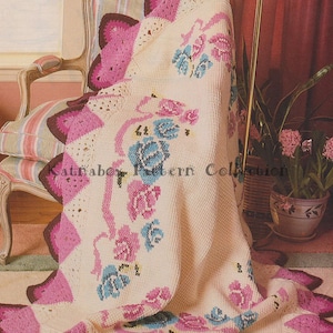 May include: A crocheted throw blanket with a scalloped edge and a floral design in shades of pink, blue, and white. The blanket is draped over a chair with a striped cushion.
