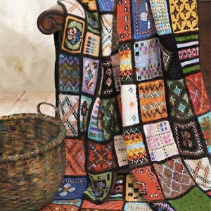 May include: A multi-colored crochet blanket composed of numerous rectangular patches featuring diverse geometric and floral patterns in shades of orange, blue, yellow, green, and white, all bordered by dark brown yarn, draped over a wooden surface.