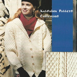 May include: A white crocheted cardigan with a cable knit pattern and a shawl collar. The cardigan has a button closure and is shown being worn by a woman with a red scarf and a black beret. The image also includes the text "Katnabox Pattern Collection".