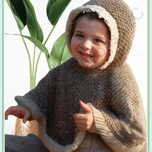 Op de afbeelding: Een kind draagt een handgemaakte bruine gebreide poncho met een capuchon en beige rand. Het kind lacht en geeft een duim omhoog. De achtergrond bevat groene bladeren.