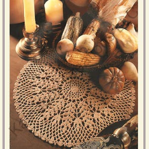 May include: A crocheted, round doily in a light brown hue, featuring an intricate openwork design. The doily is placed on a wooden surface, alongside a basket of gourds, corn, and candles, creating a warm, autumnal theme.