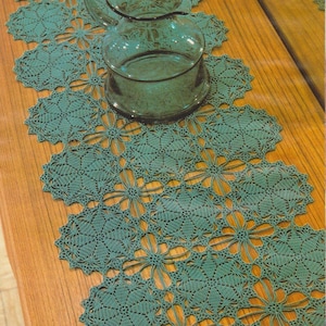 May include: A green crocheted table runner with a repeating pattern of flowers. The runner is on a wooden table with a clear glass jug on top.