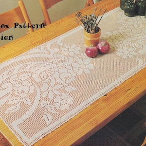 Puede incluir: Un camino de mesa de crochet blanco con un diseño floral. El camino de mesa está colocado sobre una mesa de madera con dos sillas y un jarrón de utensilios. El texto "Katnábox Pattern Collection" es visible en la esquina superior izquierda de la imagen.
