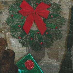May include: A green crocheted wreath with red berries and a red velvet bow. The wreath is hanging on a stone wall with a broom and wrapped gifts in the foreground.