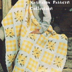 Op de afbeelding: Een geel en wit gehaakt deken met een bloemenpatroon. Het deken is gevouwen en gedrapeerd over de schoot van een vrouw. De tekst "Katrïabox Pattern Collection" is zichtbaar in de rechterbovenhoek van de afbeelding.