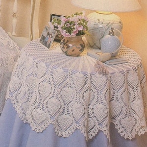 May include: A white crocheted table cloth with a delicate, lacy design. The tablecloth is laid over a light blue table and features a scalloped edge. A small bouquet of pink flowers in a brown ceramic vase sits on the table, along with a lamp and a book.