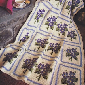 May include: A crocheted blanket with a pattern of purple flowers and green leaves. The blanket is made of white yarn with blue trim.
