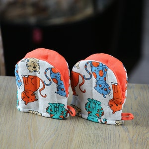 May include: A pair of orange and white oven mitts with a cat print design.