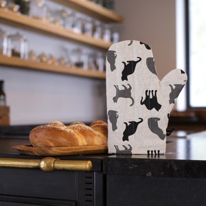 Pode incluir: Uma luva de forno preta e branca com um padrão de gato. A luva está sobre uma bancada preta ao lado de um pão em uma tábua de cortar de madeira.