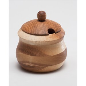 May include: A wooden sugar bowl with a lid and a knob on top. The bowl is made of light and dark wood and has a swirling pattern. The lid has a small opening for pouring.