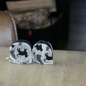 May include: A pair of black and white oven mitts with a cat silhouette pattern. The mitts are made of a soft, quilted fabric and have a loop for hanging.