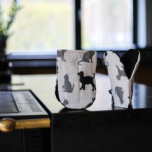 Pode incluir: Um par de luvas de forno com um design de estampa de gatos preto e branco. As luvas são feitas de um tecido claro e têm um forro preto resistente ao calor.
