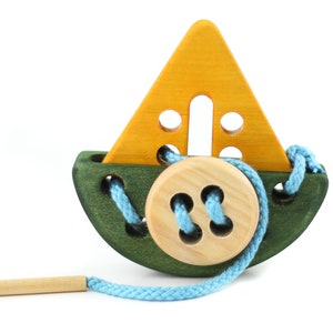 Könnte beinhalten: Ein hölzernes Spielzeugboot mit grünem Rumpf, gelbem Segel und einem Holzknopf. Eine blaue Schnur ist durch die Löcher im Boot und im Knopf gefädelt. Eine Holznadel ist im Lieferumfang enthalten.