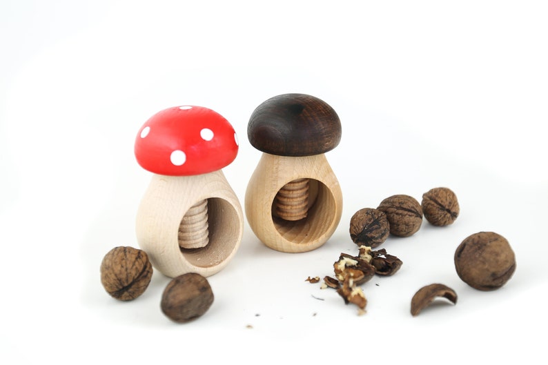 May include: Two wooden mushroom nutcrackers, one with a red and white cap and the other with a brown cap, with walnuts and walnut shells scattered around them.