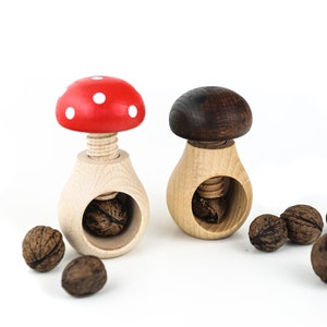 May include: Two wooden nutcrackers shaped like mushrooms. One is red with white dots and the other is brown. Both have a wooden screw to crack nuts. There are several walnuts scattered around the nutcrackers.