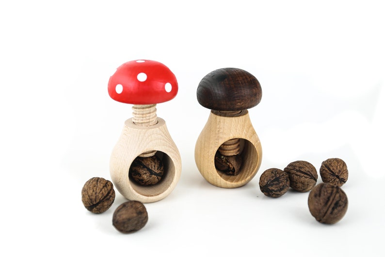 May include: Two wooden nutcrackers shaped like mushrooms. One is red with white dots and the other is brown. Both have a wooden screw to crack nuts. There are several walnuts scattered around the nutcrackers.