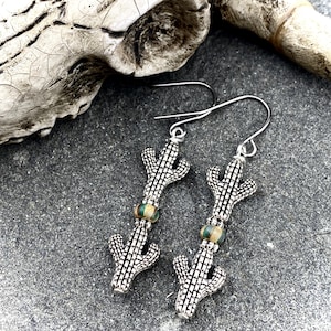 May include: Silver-toned cactus earrings with a textured design. Each earring features a cactus charm and a small, round bead with green and gold tones. The earrings hang from silver-colored ear wires.
