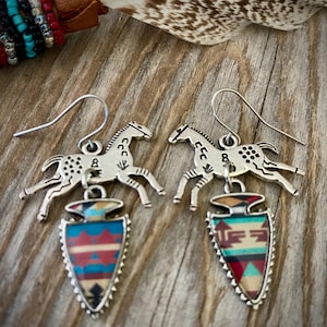May include: Silver-toned earrings featuring a horse silhouette above a colorful arrowhead pendant. The pendants display a geometric pattern in shades of red, blue, and beige. The earrings are set against a rustic wooden background.