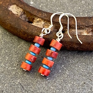 May include: A pair of handmade earrings featuring a combination of red and blue beads. The earrings have a silver hook and are composed of cylindrical red beads interspersed with small, round blue beads. The earrings are displayed on a gray surface.