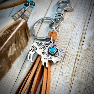 May include: A silver-colored keychain with a buffalo charm, turquoise stone, and brown leather fringe. The buffalo charm has an etched design and is attached to a silver keyring and clasp. A feather tassel with turquoise beads is also visible.