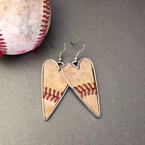 Puede incluir: Pendientes en forma de coraz&oacute;n con tem&aacute;tica de b&eacute;isbol. Los pendientes presentan un fondo beige con costuras rojas, enmarcados por un borde de metal plateado. Un bal&oacute;n de b&eacute;isbol es visible en el fondo.