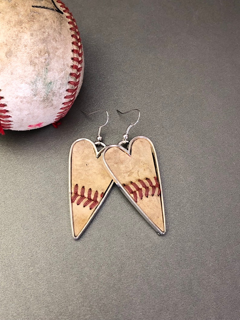 Puede incluir: Pendientes en forma de coraz&oacute;n con tem&aacute;tica de b&eacute;isbol. Los pendientes presentan un fondo beige con costuras rojas, enmarcados por un borde de metal plateado. Un bal&oacute;n de b&eacute;isbol es visible en el fondo.