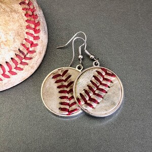 Puede incluir: Pendientes de b&eacute;isbol plateados con dise&ntilde;o de b&eacute;isbol. Los pendientes presentan un dise&ntilde;o circular con costuras rojas sobre un fondo blanco, imitando una pelota de b&eacute;isbol. Una parte de una pelota de b&eacute;isbol es visible en el fondo.