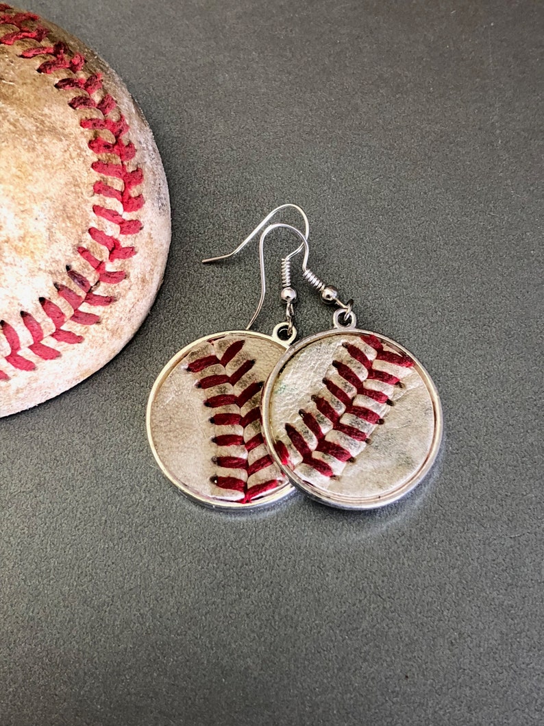 Puede incluir: Pendientes de b&eacute;isbol plateados con dise&ntilde;o de b&eacute;isbol. Los pendientes presentan un dise&ntilde;o circular con costuras rojas sobre un fondo blanco, imitando una pelota de b&eacute;isbol. Una parte de una pelota de b&eacute;isbol es visible en el fondo.