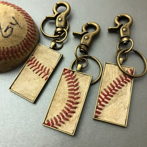 Puede incluir: Tres llaveros con tem&aacute;tica de b&eacute;isbol con herrajes de color bronce y una pelota de b&eacute;isbol. Cada llavero presenta un colgante rectangular con un dise&ntilde;o de costura de b&eacute;isbol en hilo rojo sobre un fondo beige. La pelota de b&eacute;isbol es beige con costuras rojas.