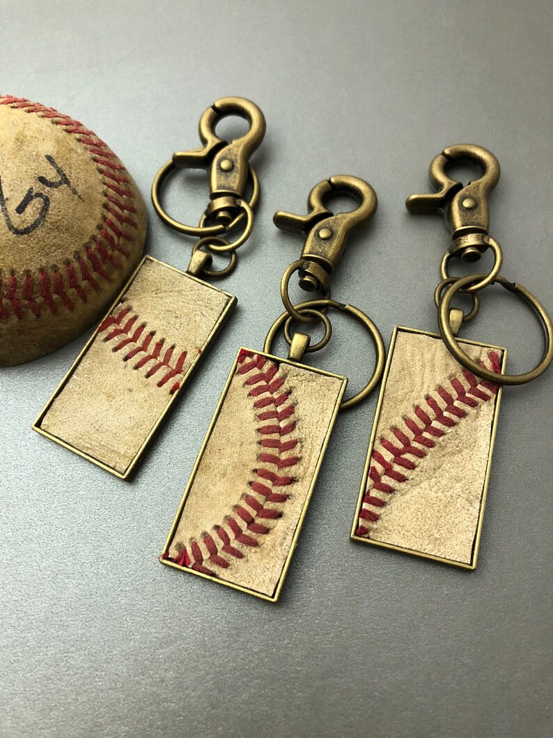 Puede incluir: Tres llaveros con tem&aacute;tica de b&eacute;isbol con herrajes de color bronce y una pelota de b&eacute;isbol. Cada llavero presenta un colgante rectangular con un dise&ntilde;o de costura de b&eacute;isbol en hilo rojo sobre un fondo beige. La pelota de b&eacute;isbol es beige con costuras rojas.
