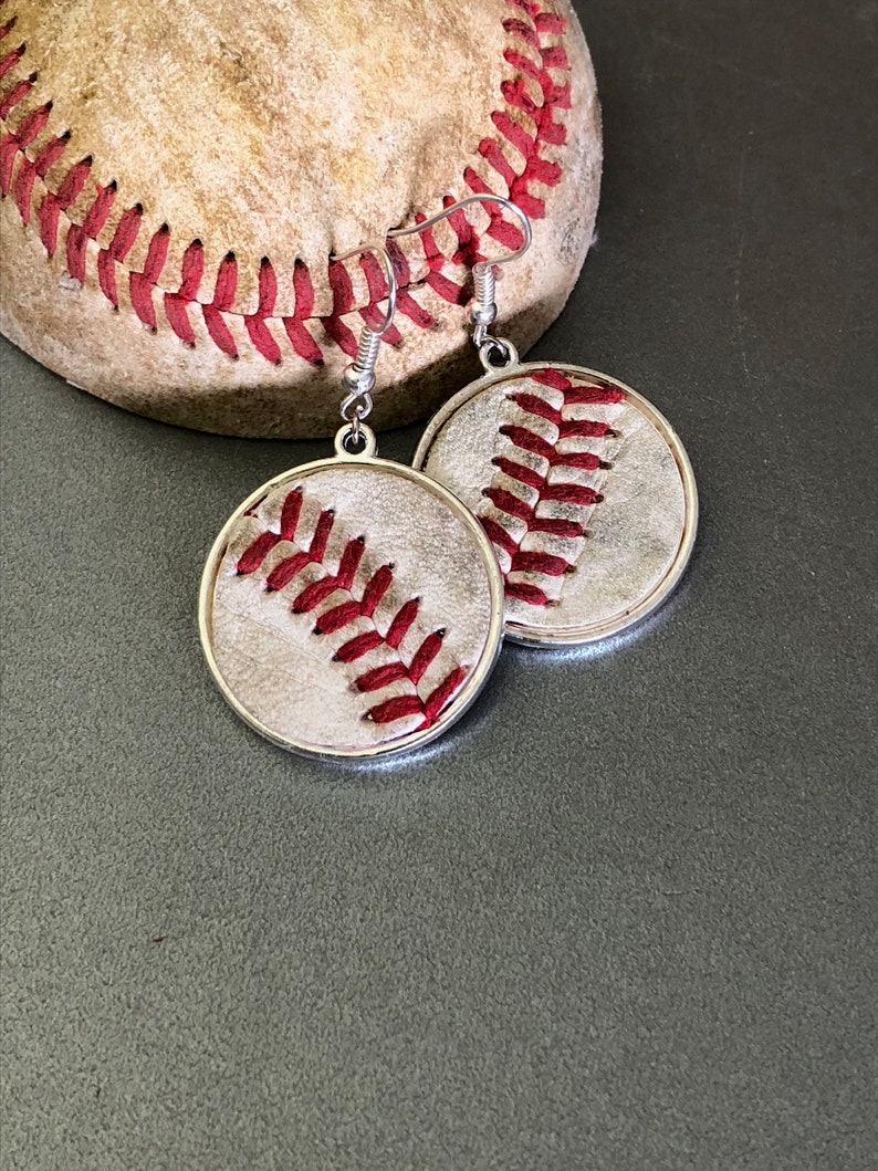 Puede incluir: Pendientes de b&eacute;isbol plateados con dise&ntilde;o de b&eacute;isbol. Los pendientes tienen un colgante circular con un dise&ntilde;o de costura de b&eacute;isbol en hilo rojo. Un bal&oacute;n de b&eacute;isbol est&aacute; en el fondo. Para los fan&aacute;ticos del deporte.
