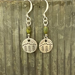May include: Silver earrings with a mountain and pine tree design. Each earring features a green stone bead.