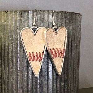 Puede incluir: Pendientes en forma de coraz&oacute;n con tem&aacute;tica de b&eacute;isbol. Los pendientes presentan un fondo beige claro con costuras rojas, enmarcados por un borde de metal plateado. Cuelgan de ganchos plateados, ofreciendo un accesorio &uacute;nico.